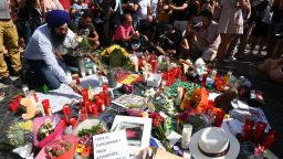 People stand next to flowers, candles and other items set up on the Las Ramblas boulevard in Barcelona as they pay tribute to the victims of the Barcelona attack, a day after a van ploughed into the crowd, killing 13 persons and injuring over 100 on August 18, 2017. 