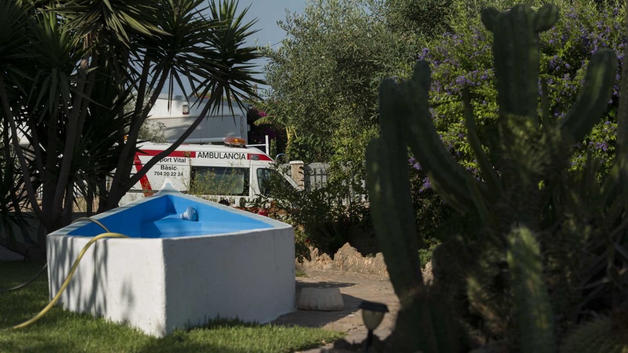 An ambulance is seen through a neighbor's front lawn, where a hose dangles in a half-empty pool.
