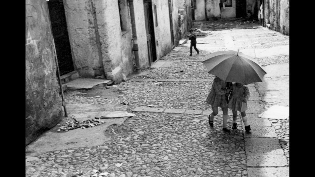 Two little girls take shelter under an umbrella in a working-class area of Palermo while a boy, whose legs are almost mirror images of those of one of the girls, walks behind them. 