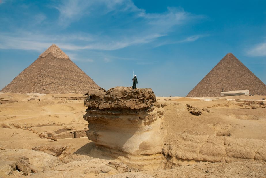 A man stands on the original surface level of the Giza plateau. Around it rock had been quarried for use on the surrounding structures. In the background, two of Giza's 4th dynasty pyramids. The Great Pyramid of Khufu covers an area of 5.3 hectares and was built with approximately 2.3 million stone blocks. <br />