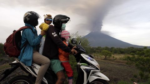 A family on a motorcycle passes by the Mount Agung volcano erupting in the background.