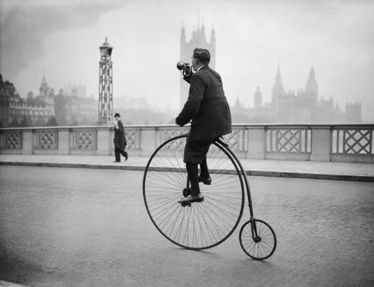 Lambeth Bridge in 1932, featuring a gentleman riding a penny farthing bicycle, he's blowing a bugle to warn people he's coming. 