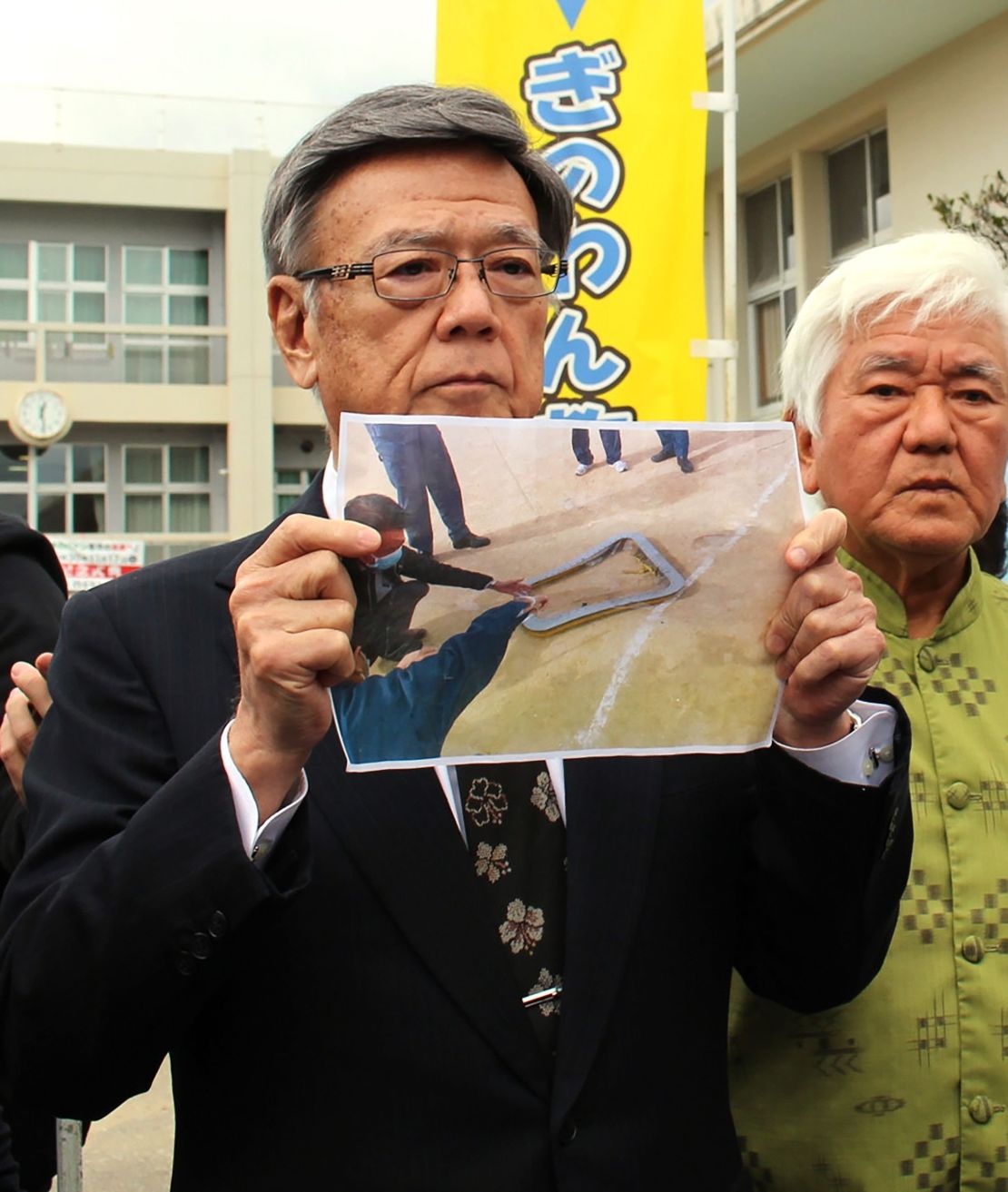 Okinawa Governor Takeshi Onaga stands outside an elementary school as he shows a picture of the window that fell from a US military helicopter.