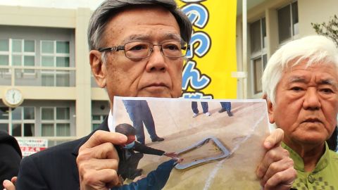 Okinawa Governor Takeshi Onaga stands outside an elementary school as he shows a picture of the window that fell from a US military helicopter.