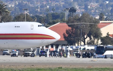 A team in white biohazard suits watch as some of the approximately 200 passengers walk to waiting buses upon arriving on a charter flight from Wuhan, China, after landing at March Air Reserve Base in Riverside, California, on Wednesday, January 29.