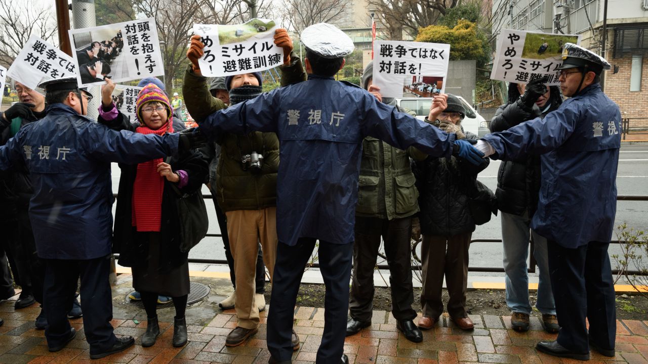 Protesters hold signs as police officers form a cordon during Tokyo's missile-attack drill Monday.