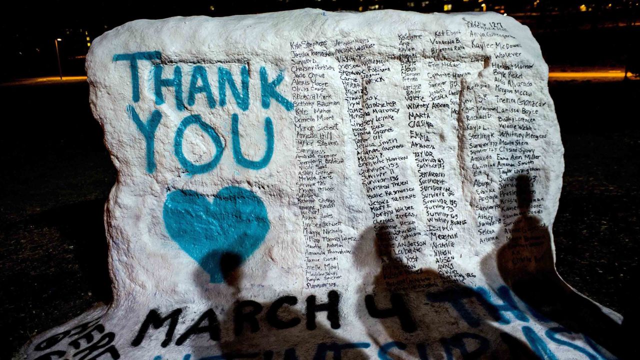 Shadows of Michigan State University students fall on "The Rock" as they examine its new, hand-painted tribute to Larry Nassar's victims.