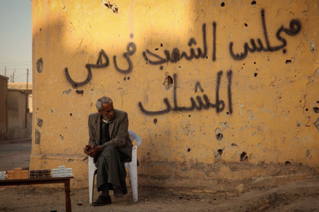 A man sitting under a graffiti that says "the people's council in Al-Mashlib neighbourhood" at the side of a street in Al-Mashlib neighbourhood in Raqqa city, Syria. 