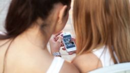 Two cute beautiful young women friends sitting on park bench on summer day, browsing pictures on smartphone, back view, focus on device screen; Shutterstock ID 295469396; Job: -