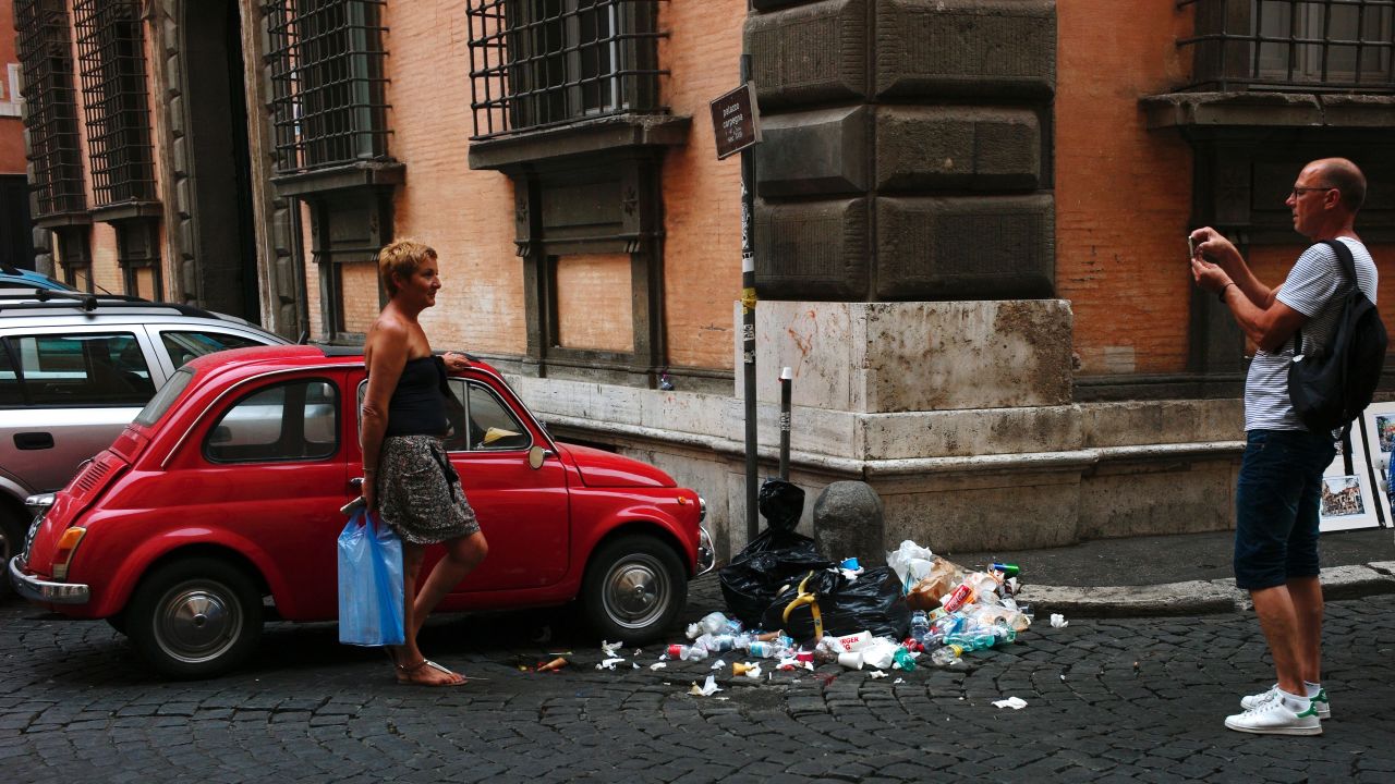 A man takes a picture of a woman standing next to an old Fiat 500 car parked in front of a pile of rubbish in a street of central Rome on July 23, 2016. (AFP / FILIPPO MONTEFORTE)