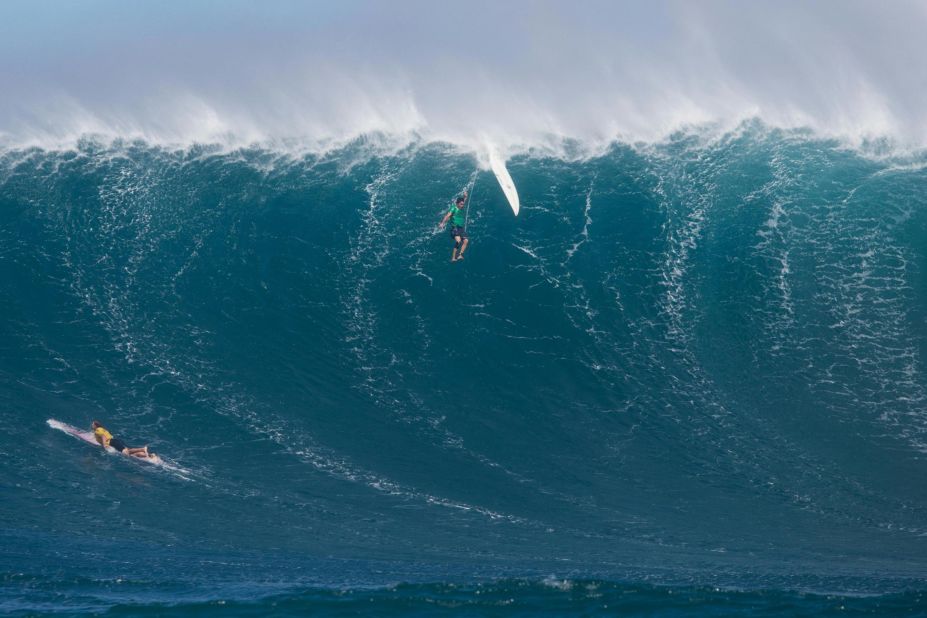 Another surfer is eaten up by a big wave as another one paddles to escape it.