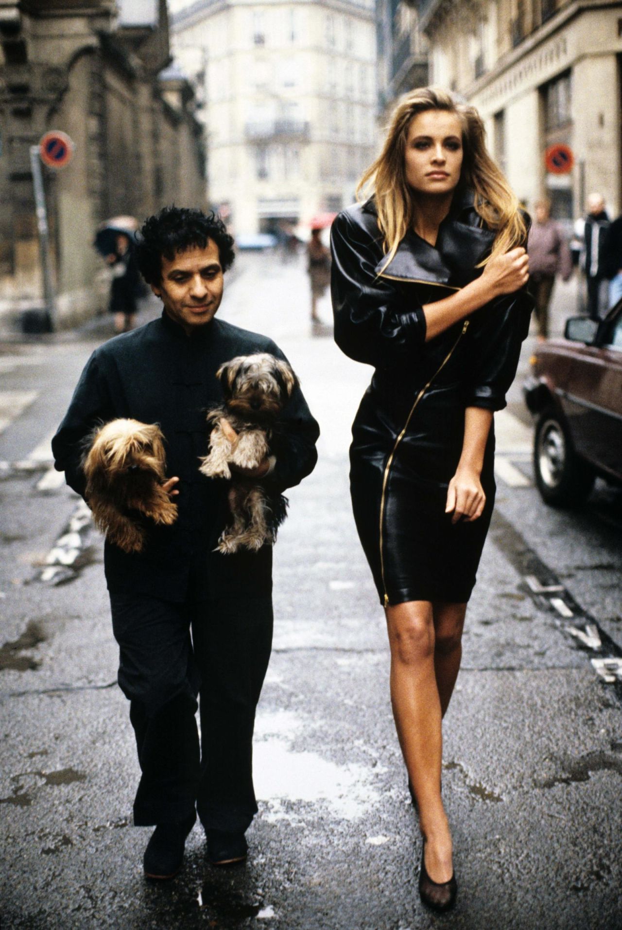 Azzedine Alaïa, holding his Yorkshire terriers, Patapouf and Wabo, walks the streets of Paris with model Frederique, who wears a dress of his design.