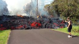 In this photo taken from video an unidentified man gets close to a lava flow advancing down a road in the Leilani Estates subdivision near Pahoa on the island of Hawaii Monday, May 7, 2018. Kilauea volcano has destroyed more than two dozen homes since it began spewing lava hundreds of feet into the air last week, and residents who evacuated don't know how long they might be displaced. The decimated homes were in the Leilani Estates subdivision, where molten rock, toxic gas and steam have been bursting through openings in the ground created by the volcano. (Scott Wiggers/Apau Hawaii Tours via AP)