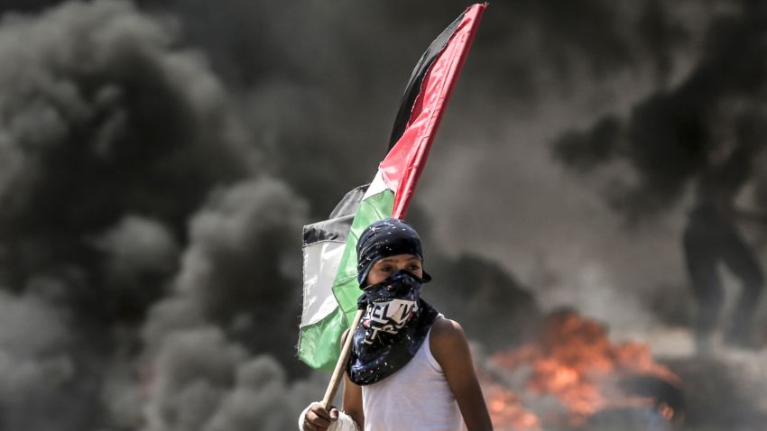 TOPSHOT - A Palestinian boy holding his national flag looks at clashes with Israeli security forces near the border between the Gaza Strip and Israel east of Gaza City on May 14, 2018, as Palestinians protest over the inauguration of the US embassy following its controversial move to Jerusalem. - Dozens of Palestinians were killed by Israeli fire on May 14 as tens of thousands protested and clashes erupted along the Gaza border against the US transfer of its embassy to Jerusalem, after months of global outcry, Palestinian anger and exuberant praise from Israelis over President Donald Trump's decision tossing aside decades of precedent. (Photo by MAHMUD HAMS / AFP)        (Photo credit should read MAHMUD HAMS/AFP/Getty Images)