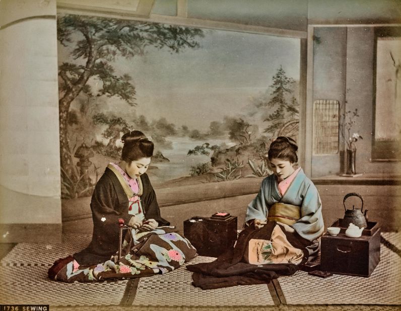 A photo shows two women sewing in a traditional setting. Traditional furnishing elements such as portable brazier chest with the kettle and painted scroll are displayed around them.
