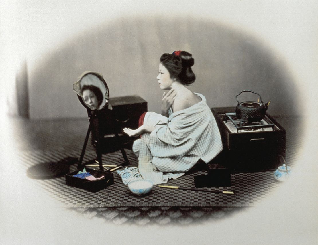 This photo, enclosed in Beato's signature oval frame, shows a woman applying white makeup to her face while leaving one shoulder uncovered. 