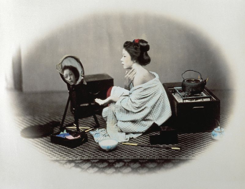 A women is applying white makeup to her face leaving one shoulder uncovered, a sensual image that attracted a lot of travelers in Japan at the end of the 19th century. 