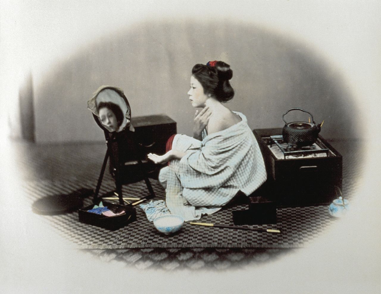 This photo, enclosed in Beato's signature oval frame, shows a woman applying white makeup to her face while leaving one shoulder uncovered. 