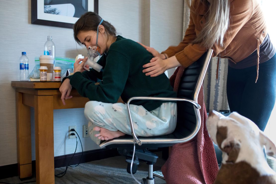 Claire breathes in medicine through a nebulizer as Larissa pounds her back with cupped hands to help loosen mucus in her airways. 
