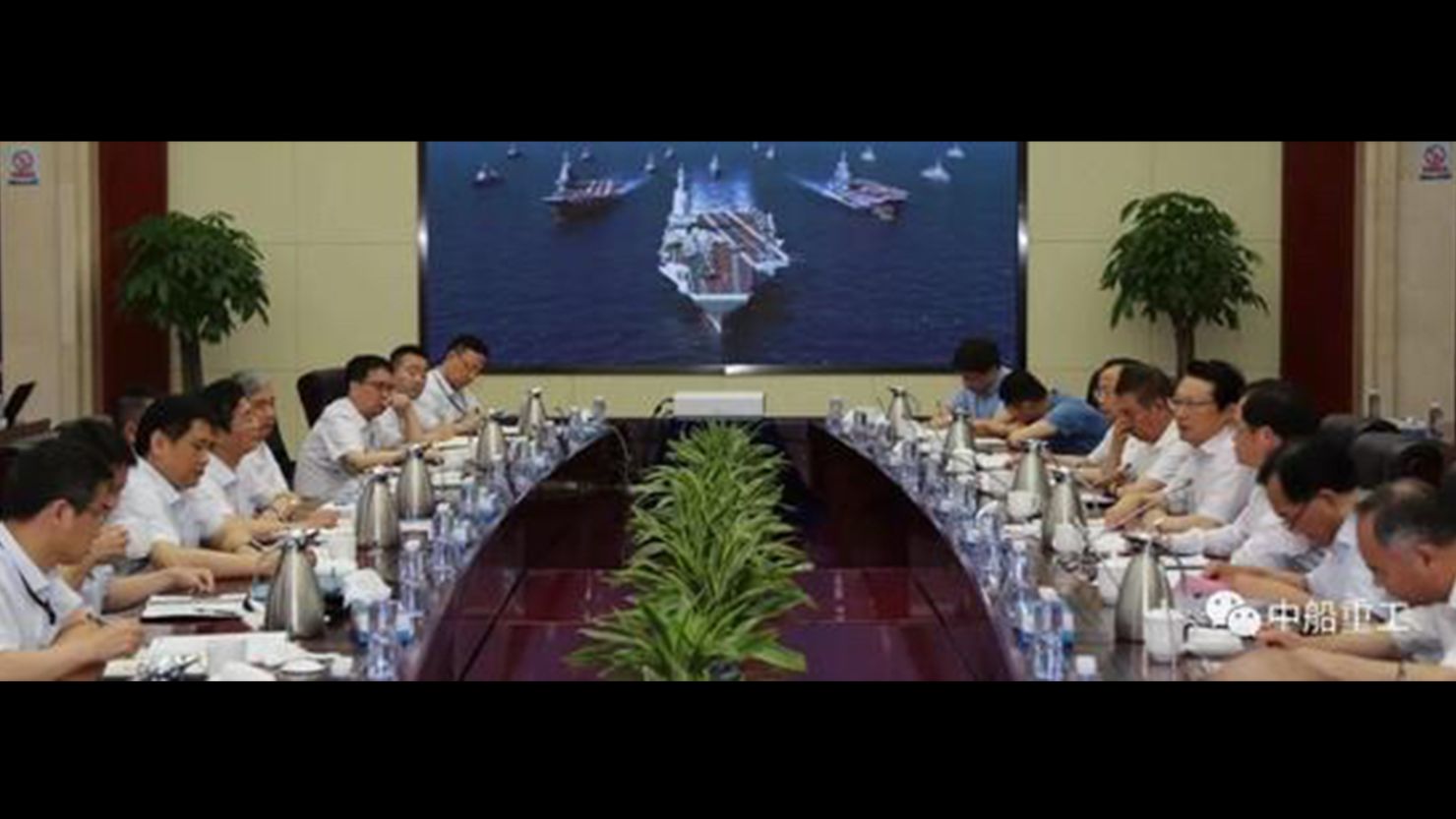 Screen shot from Chinese media shows a conference at the No.701 Research Institute of China Shipbuilding Industry Corporation with picture in the background of China's two current ski-jump carriers flanking a larger ship with a flat deck thought to picture what China's third aircraft carrier will look like.
