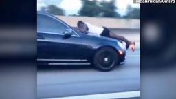 Man clinging to hood of car doing 70 mph calls 911. CNN's Jeanne Moos has the tapes.       Hood Hanger 911   Maybe you've seen the amazing vid of the guy clinging to the hood of a Mercedes doing 70 mph on the interstate in Miami. He was using one hand to hang on and holding the phone in the other. It went viral but no one knew the whole story till now. It's a doozy. While clinging to the car the guy called 911 ("I'm on top of a speeding vehicle! OK, what's the address?"). Turns out the woman driving was his ex from a rocky relationship. They argued over who was going to use the car and when she got in to go pick up her daughter, he jumped on the hood. She drove 15 miles (!!!!!) down the interstate with him hanging on for dear life. Eventually she pulled over (she shot video of his face through the windshield yelling "get off of my car" and another motorist captured the vid of the guy hanging on from the neighboring lane). Police came and arrested her, charged her with culpable negligence. We have sound with both the woman and the guy. She says he's an ex who won't leave her alone. His final bite..."just watch yourself and be careful who you  love." I did a pretty unusual standup lying on the hood of a crew car while the photog shot me from inside the car.  We did not drive during the standup but Time Warner security came up to say they'd gotten 5 calls from concerned citizens.