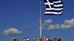 Tourists sit under a Greek flag atop Acropolis hill in Athens on August 22, 2010.  AFP PHOTO/ LOUISA GOULIAMAKI / AFP PHOTO / LOUISA GOULIAMAKI        (Photo credit should read LOUISA GOULIAMAKI/AFP/Getty Images)