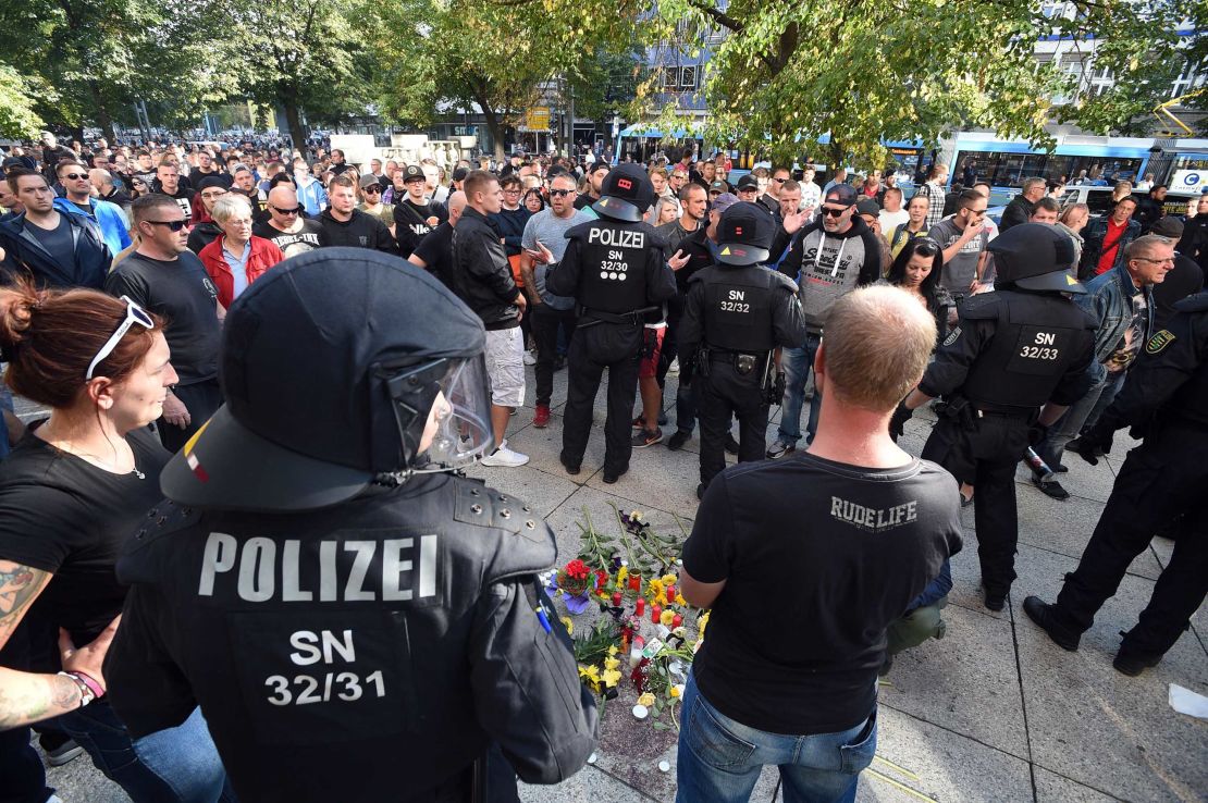 Riot police and citizens stand next to a makeshift memorial marking the site where three people were injured in a fight early Sunday. One man later died from his injuries, police said.