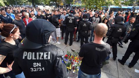Riot police and citizens stand next to a makeshift memorial marking the site where three people were injured in a fight early Sunday. One man later died from his injuries, police said.