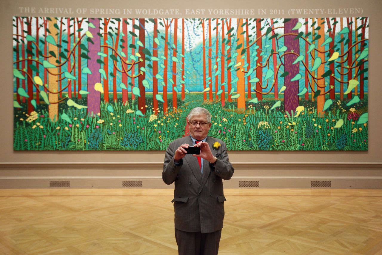British artist David Hockney takes a picture of press photographers with his mobile phone as he poses in front of his painting entitled "The Arrival of Spring in Woldgate, East Yorkshire in 2011" at the opening of his exhibition David Hockney RA: A Bigger Picture in the Royal Academy of Arts.