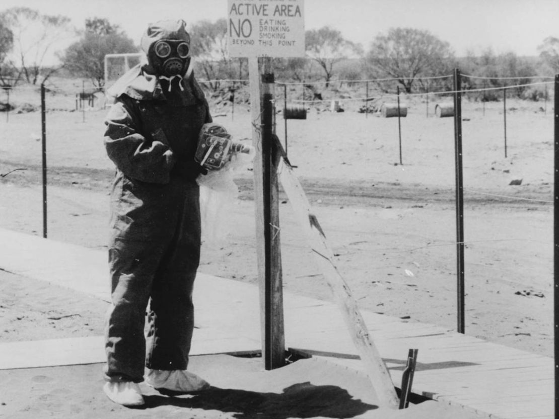 A man in protective clothing at Maralinga with a camera also protected by a plastic cover. National Archives of Australia, A6457, P214
