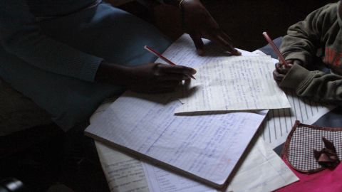 Elifuraha's son watches, pencil in hand, as she flips through her notes from a tailoring class at the Faraja Center.
