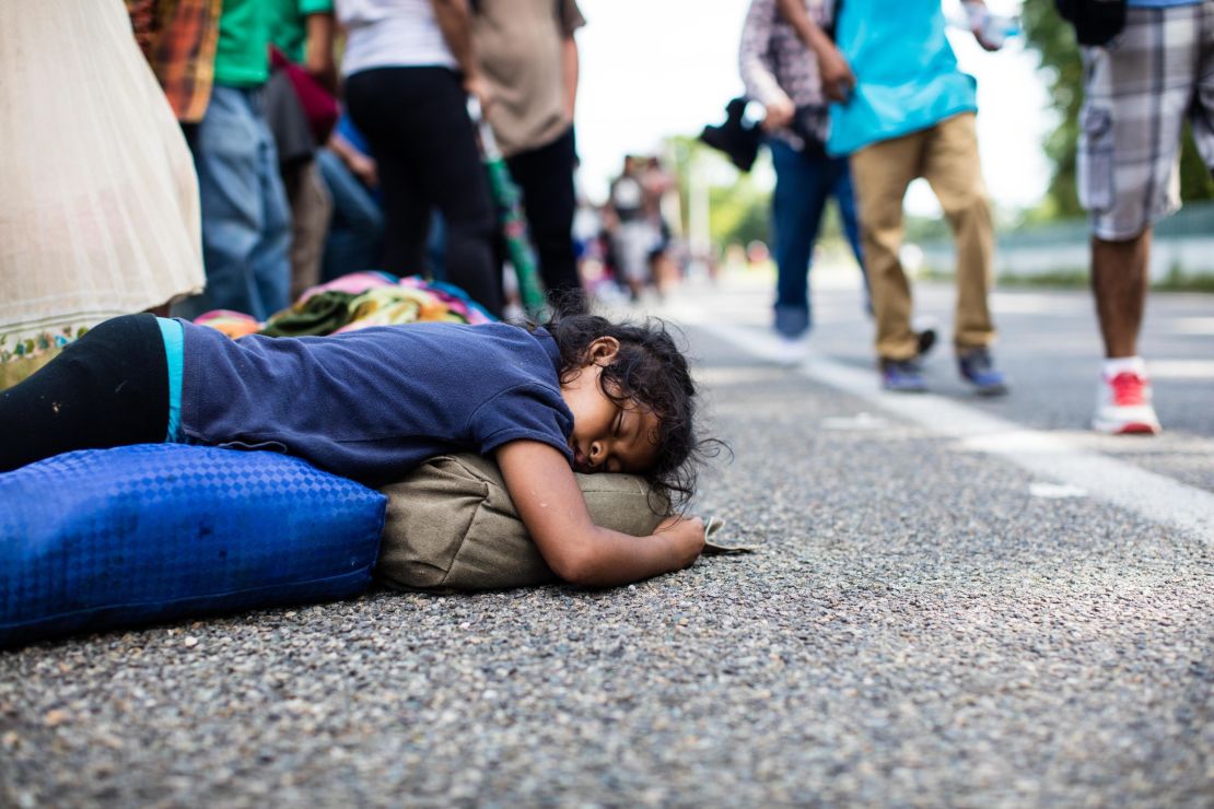 A girl sleeps on the side of the road as fellow migrants walk past.