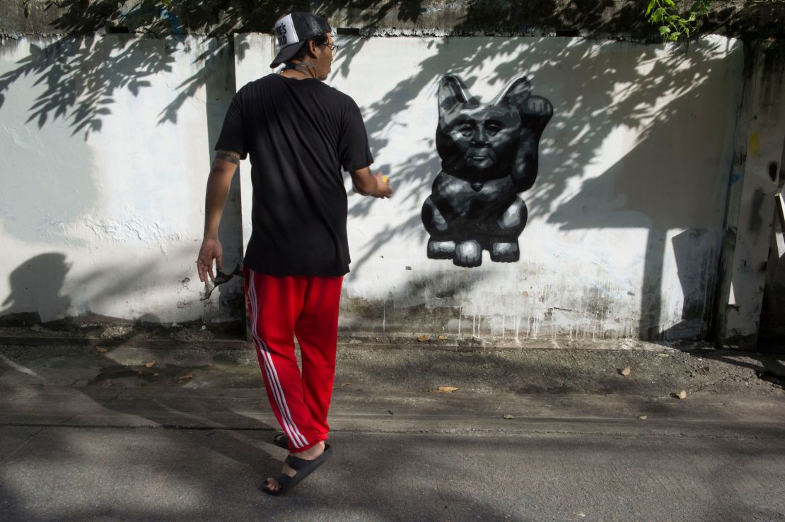 The Thai street artist who goes by the name of "Headache Stencil" walks next to his graffiti caricature of Thailand's junta chief depicted as a "lucky cat" with paw raised to rake in money, spray-painted on a fence in Bangkok. 