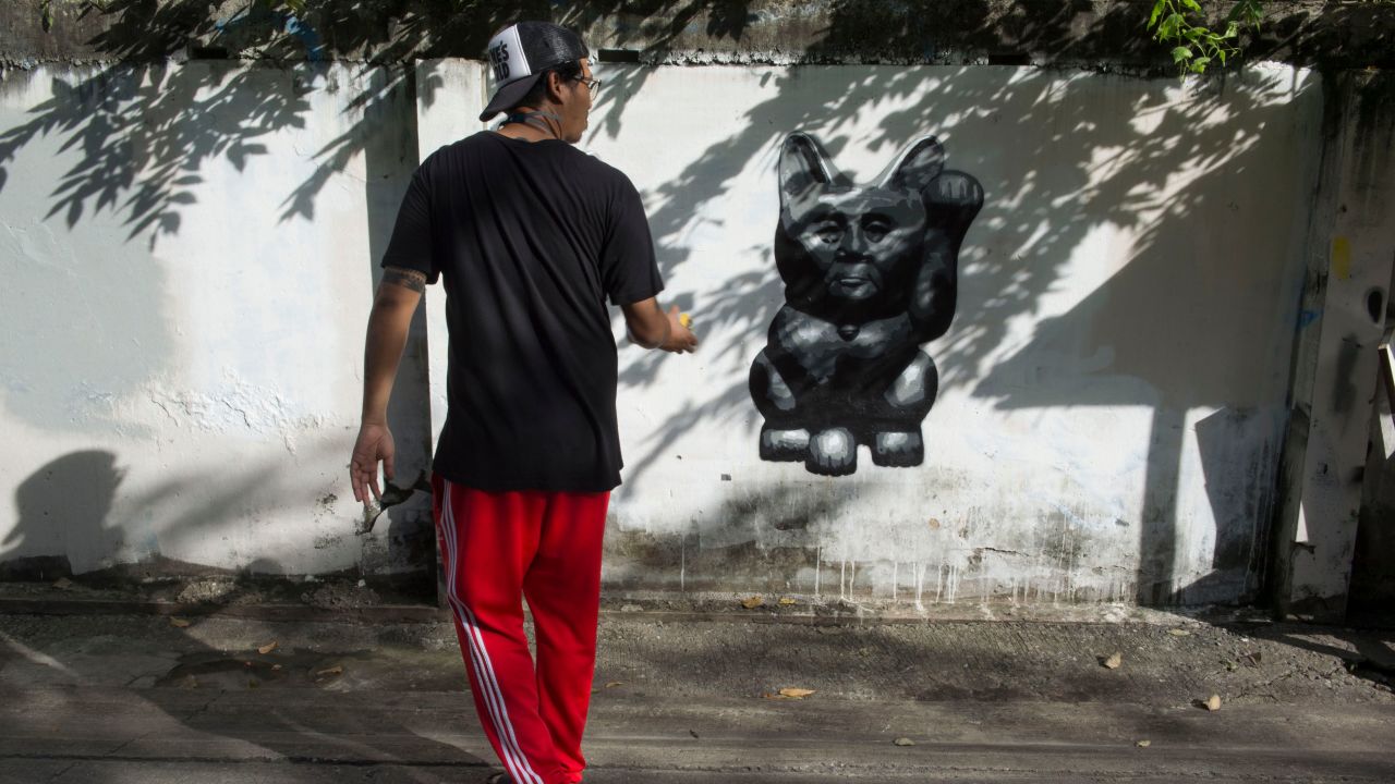 The Thai street artist who goes by the name of "Headache Stencil" walks next to his graffiti caricature of Thailand's junta chief depicted as a "lucky cat" with paw raised to rake in money, spray-painted on a fence in Bangkok. 