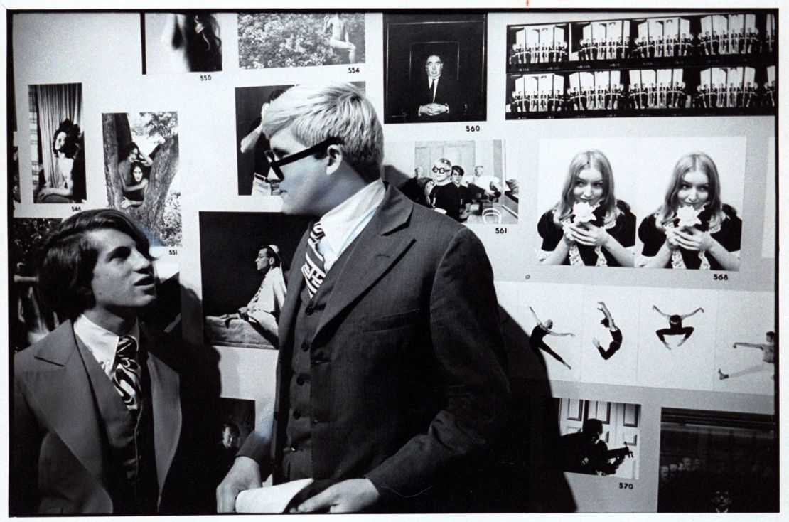 Peter Schlesinger and artist David Hockney standing in front of a photograph of themselves at a 1969 exhibition in London.