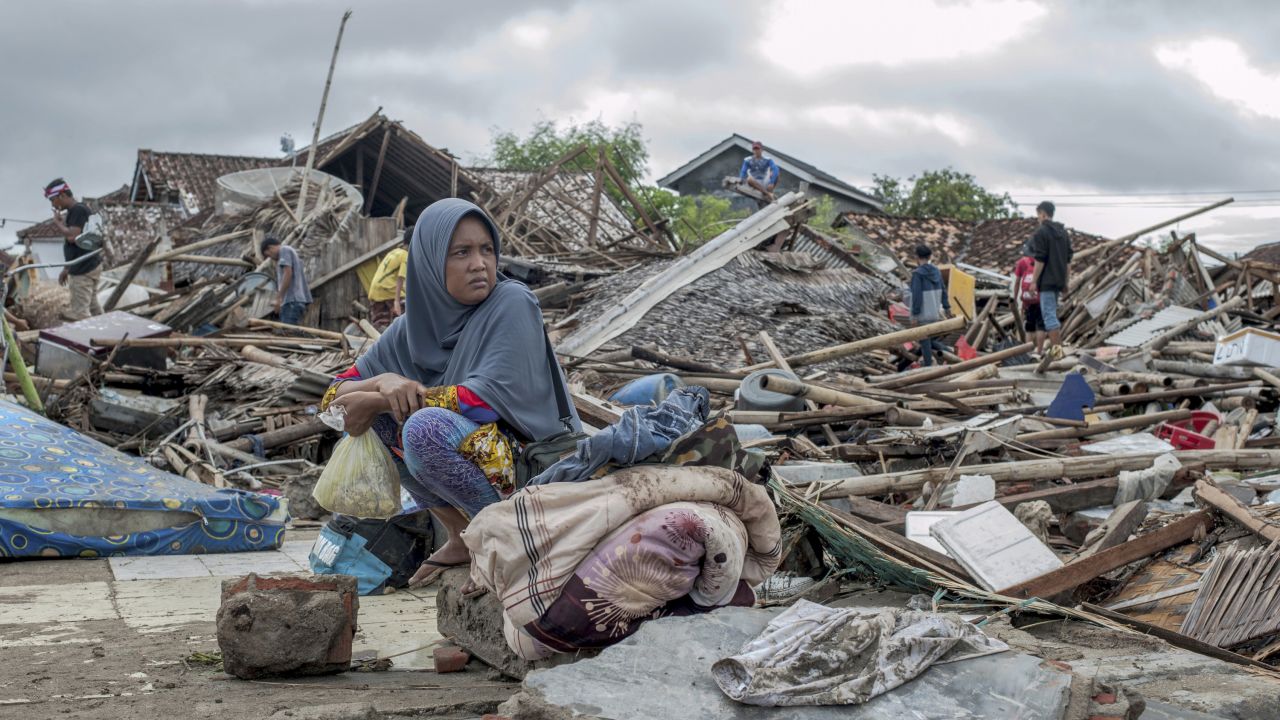 A survivor sits on debris as she salvages items from her destroyed house in Sumur, Indonesia.