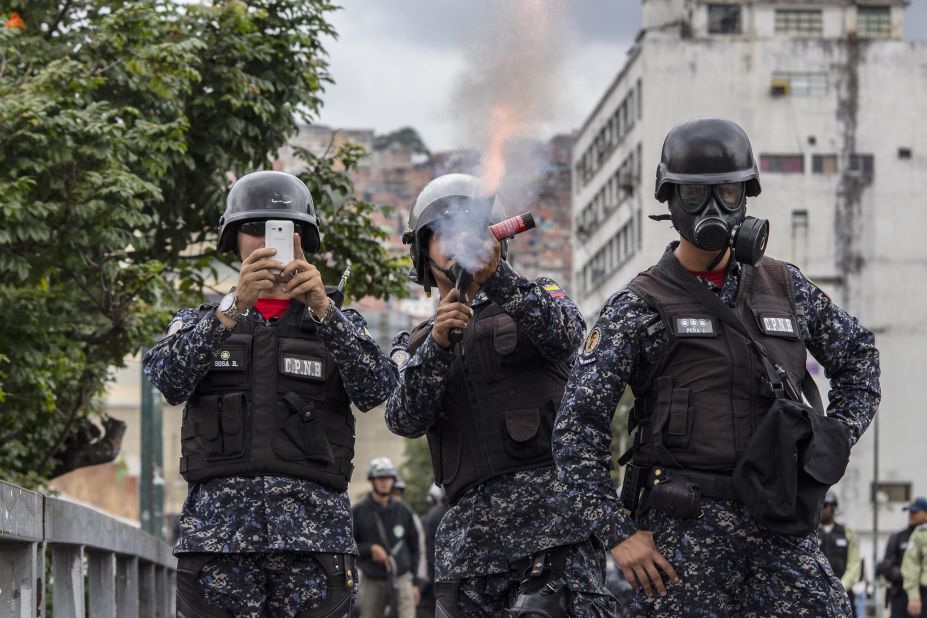 A National Police officer fires tear gas at demonstrators in Caracas while another shoots the scene with a cell phone.