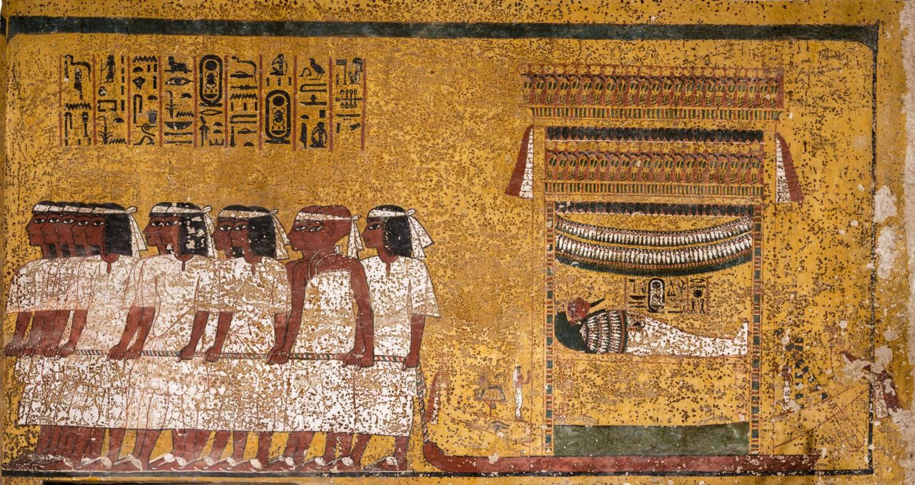 East wall of the tomb's burial chamber. Tutankhamun's mummy is shown, lying in a shrine mounted on a sledge, being drawn by 12 men in ﬁve groups. The men wear white mourning bands over their brows. The last pair, distinguished by their shaven heads and different dress, are the two viziers of Upper and Lower Egypt.