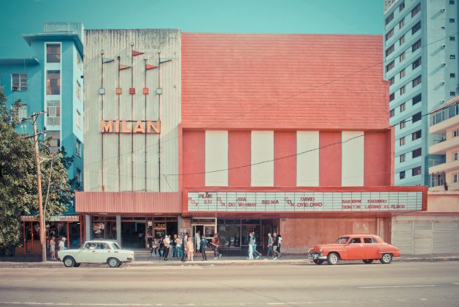 Two vintage cars sit outside a crumbling movie theater.