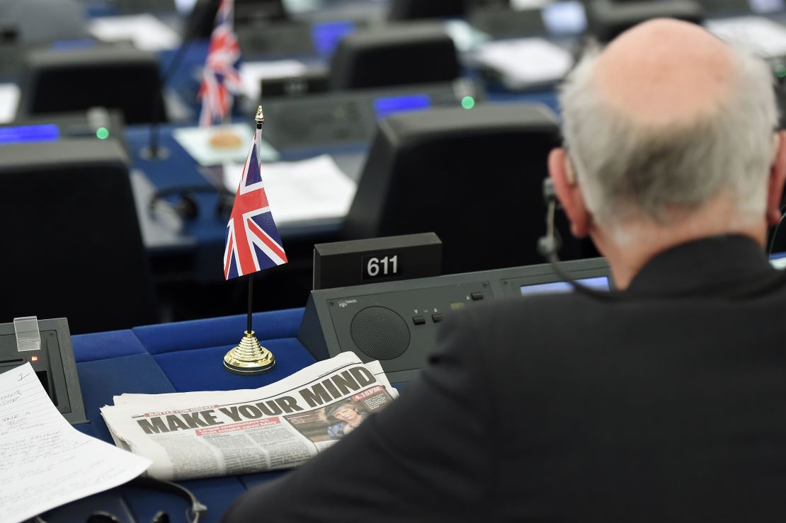 A newspaper with a Brexit-focused headline is seen next to a MEP attending a debate in Parliament.