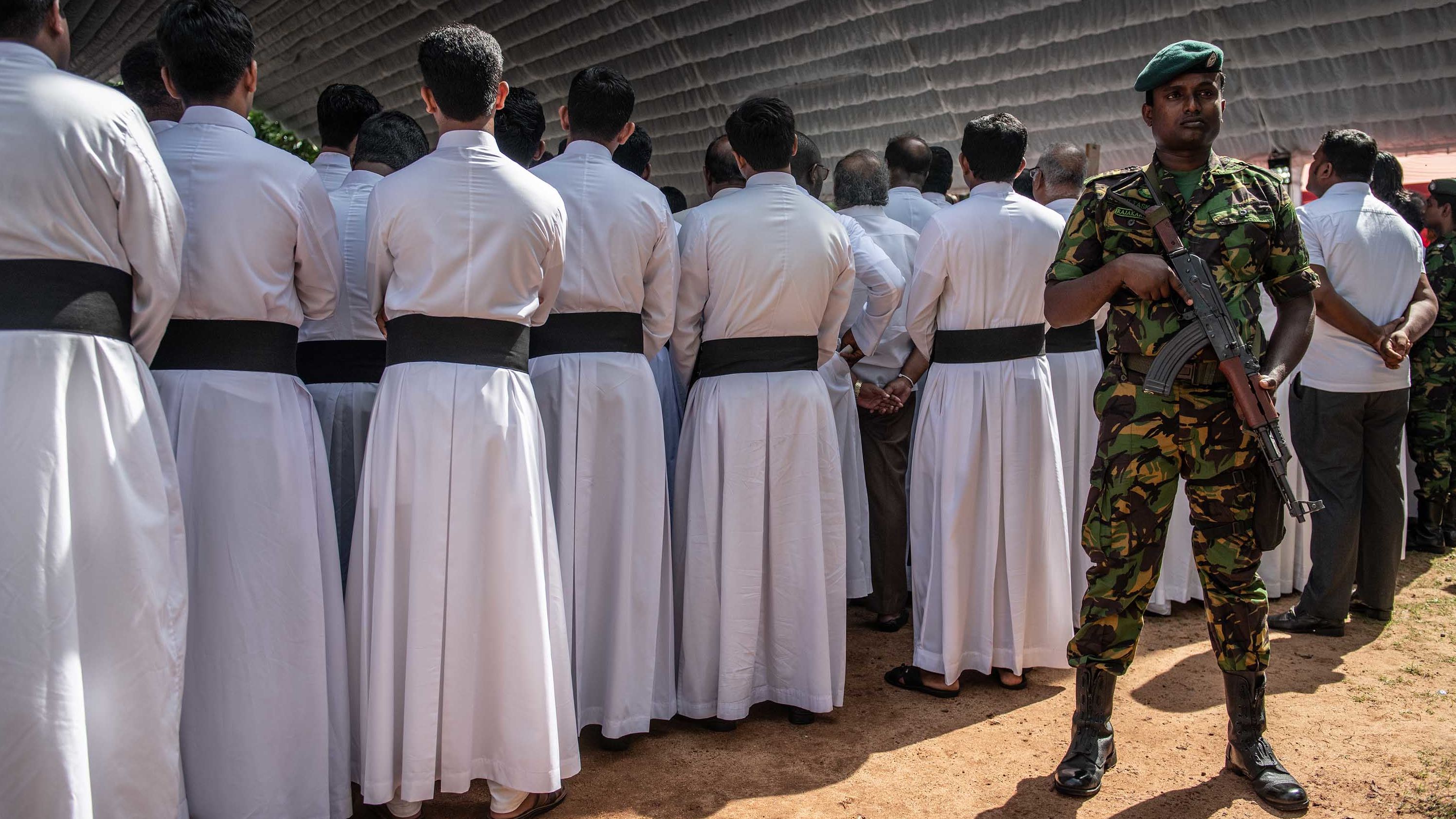 A soldier stands guard next to members of the clergy during a mass funeral in Negombo on Tuesday.
