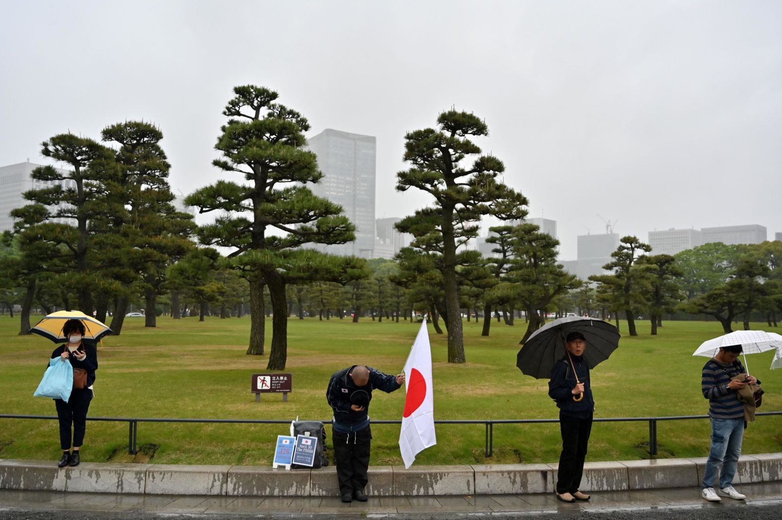 A man holds a Japanese flag and bows while people gather outside the Imperial Palace.