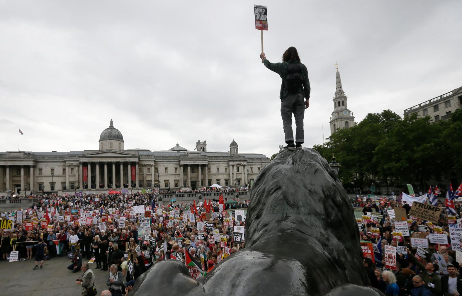 A man holds a sign reading "Dump Trump" as he stands atop a statue in Trafalgar Square.