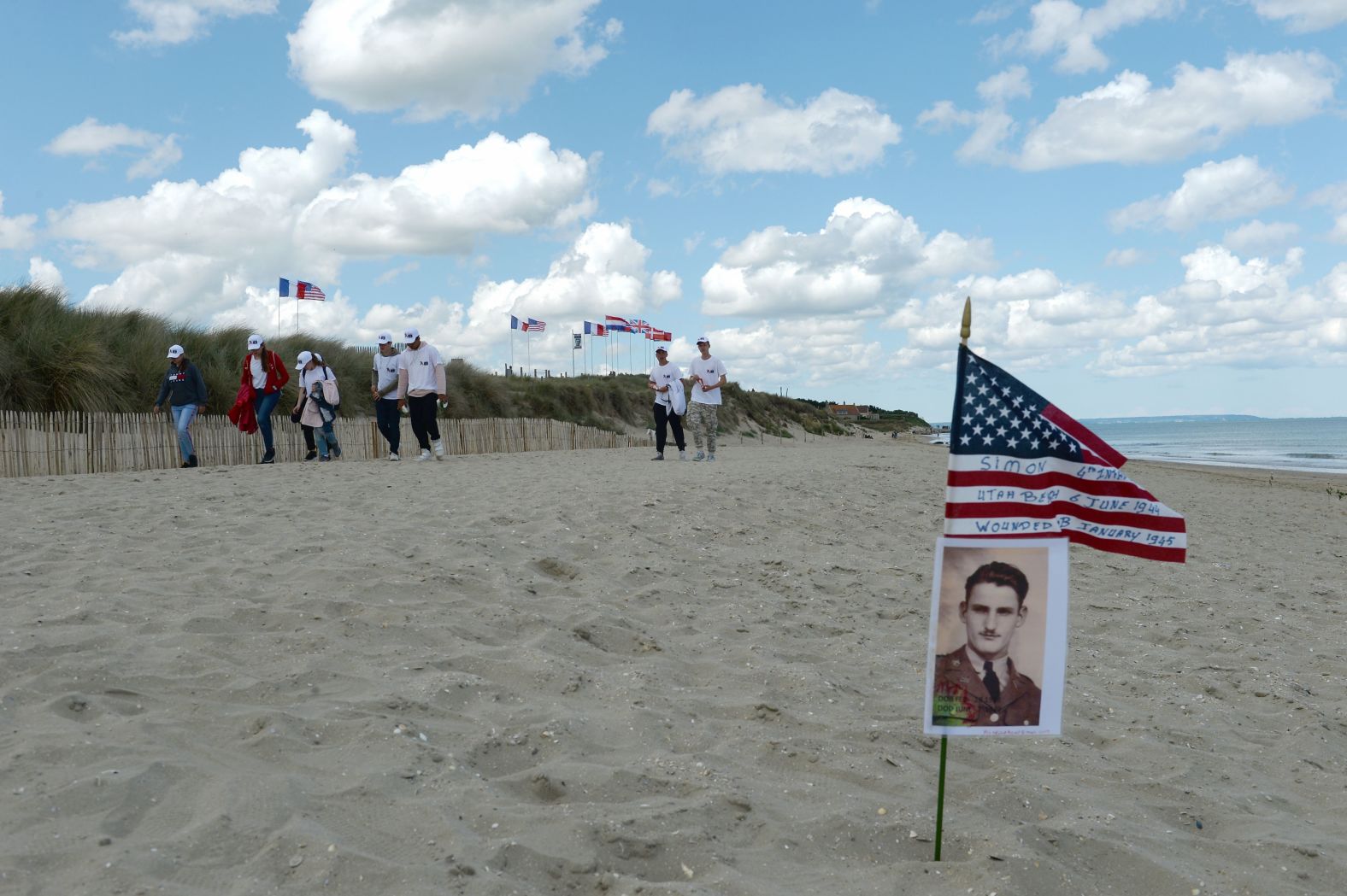 WWII enthusiasts walk by a portrait and an American flag planted in the sand at Utah Beach in Sainte-Marie-du-Mont, France. It reads: "In memory staff Sgt. Charles Franck Simon 4th Infantry division -- Utah Beach, 6 June 1944 -- Wounded."