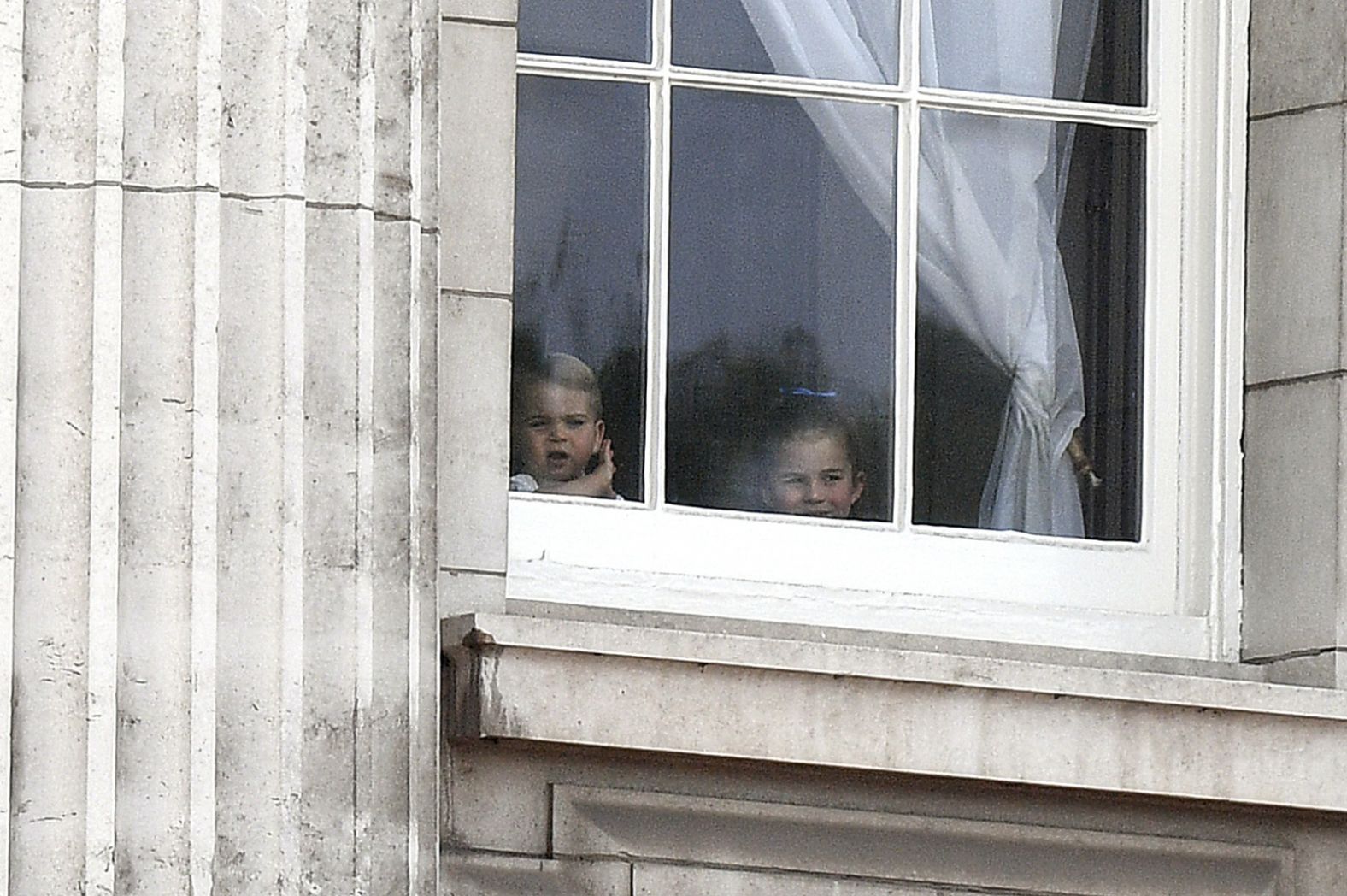 Prince Louis and Princess Charlotte gaze out a window at Buckingham Place to see soldiers on the Mall as part of the Trooping the Colour celebration. They are the children of Prince William and Catherine, Duchess of Cambridge.