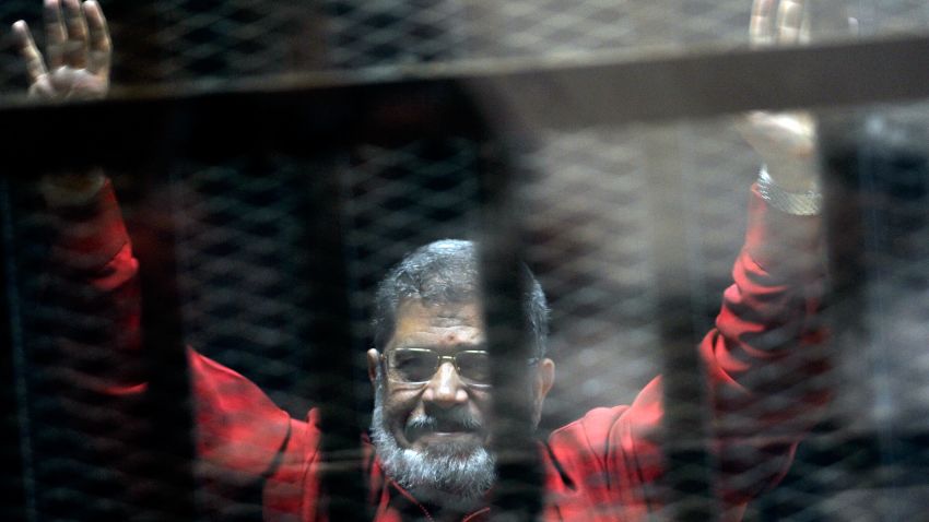 FILE - In this June 21, 2015 file photo, former Egyptian President Mohammed Morsi, wearing a red jumpsuit that designates he has been sentenced to death, raises his hands inside a defendants cage in a makeshift courtroom at the national police academy, in an eastern suburb of Cairo, Egypt. An Egyptian court has sentenced six people, including two Al-Jazeera employees, to death for allegedly passing documents related to national security to Qatar and the Doha-based TV network during the rule of Islamist president Mohammed Morsi. Morsi, the case's top defendant, was also sentenced on Saturday to 25 years in prison. He was ousted by the military in July 2013, and has already been sentenced to death in other cases. (AP Photo/Ahmed Omar, File)