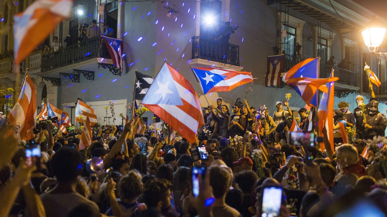 People in the crowd hold flowers and flags outside the governor's mansion.