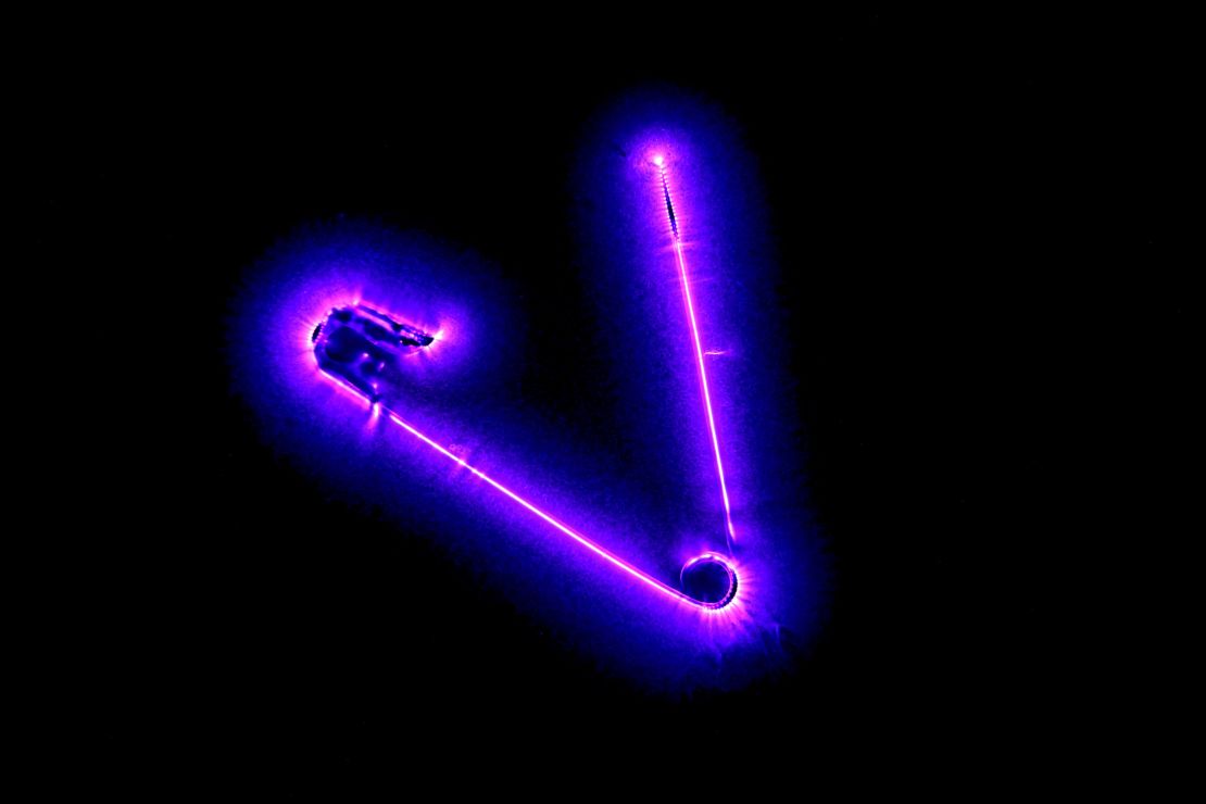 Safety Corona by Richard Germain. A safety pin is connected to a high tension AC generator. The pin ionizes the air around it. When the electrons fall back on an atom, the excess energy is emitted as a photon, which generate the corona glow around the pin. The fuzziness of the pin is because the camera did not actually capture light reflected on the pin but rather the light emitted by the ionized light around it.