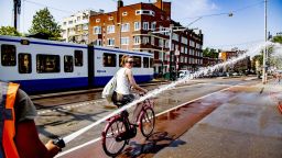A firefighter sprays water as a woman rides her bicycle on the Wiegbrug bridge, during a heatwave in Amsterdam on July 25, 2019. - On July 24 and 25, 2019, as Europe is hit by a second heatwave in less than a month, Germany, Belgium and The Netherlands top their previous heat records. The Royal Netherlands Meteorological Institute (KNMI) has issued an official warning due to the heat, advising residents of the capital city to stay inside. (Photo by Robin UTRECHT / ANP / AFP) / Netherlands OUT        (Photo credit should read ROBIN UTRECHT/AFP/Getty Images)