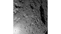 A segunda imagem da câmera MASCAM desenvolvida por DLR é direcionada obliquamente para baixo no asteroide Ryugu e cobre áreas a leste da rota de descida.  Comparado com a primeira imagem, fica claro que MASCOT se moveu de forma turbulenta em direção a Ryugu, como esperado, realizando assim curvas e capotamentos.  As imagens mostram uma pedra enorme, que ocupa a borda leste (direita) da imagem e tem várias dezenas de metros de comprimento.  No canto inferior esquerdo está a sombra do MASCOT, que o Sol atrás da sonda de pouso está projetando na superfície do asteroide: o MASCOT tem 30 centímetros de comprimento.  Ryugu é um corpo sem atmosfera, então os contornos do MASCOT são nítidos nas sombras projetadas na superfície do asteróide.
